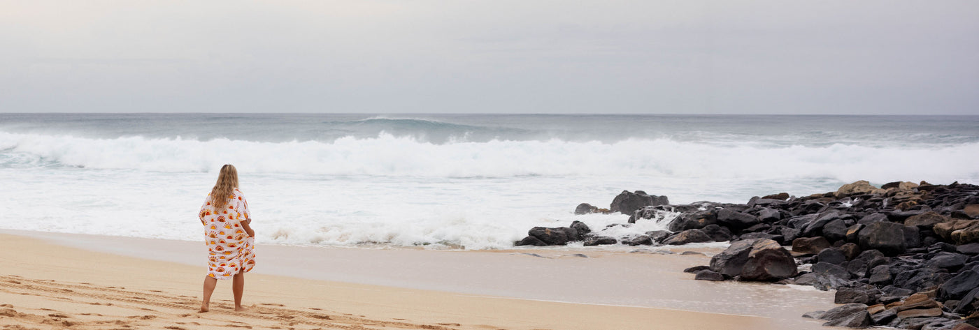 Woman in a sun patterned towel poncho standing on a beach with waves and rocks in the background