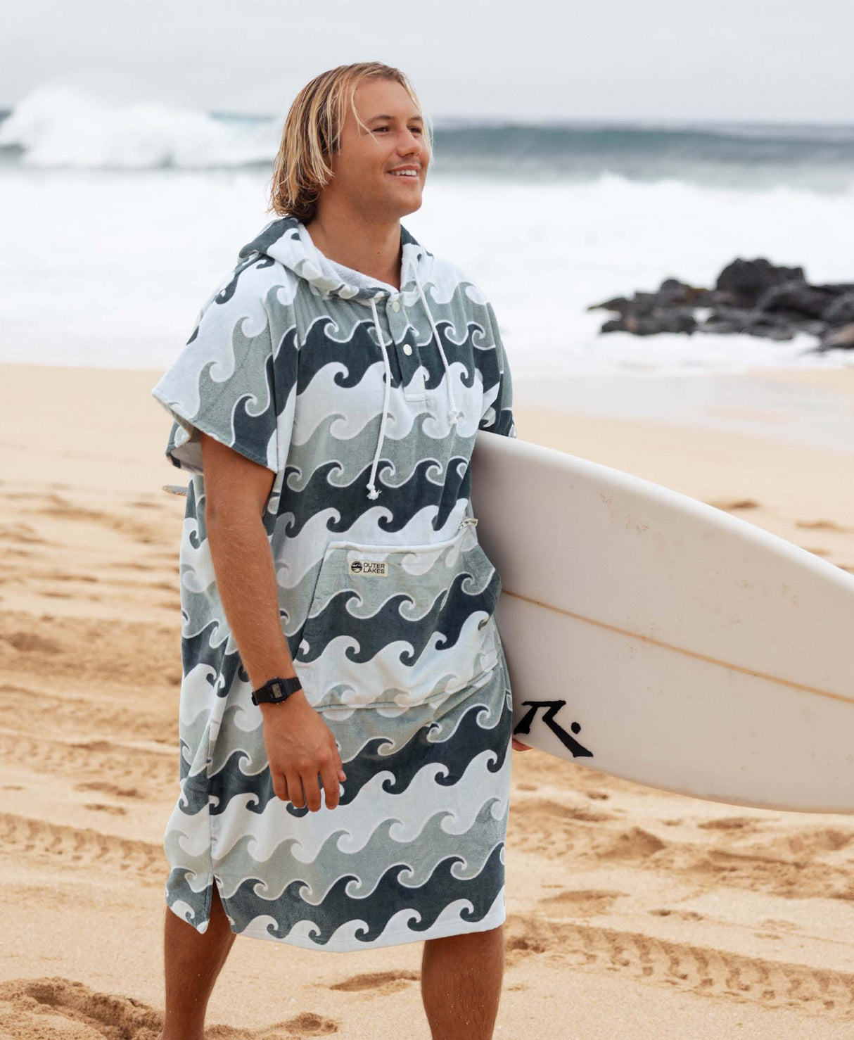 Man wearing a wave-patterned towel poncho on a beach with a surfboard.