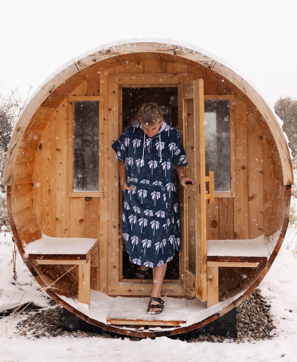 Person in a palm tree towel poncho standing in a wooden barrel sauna during winter.