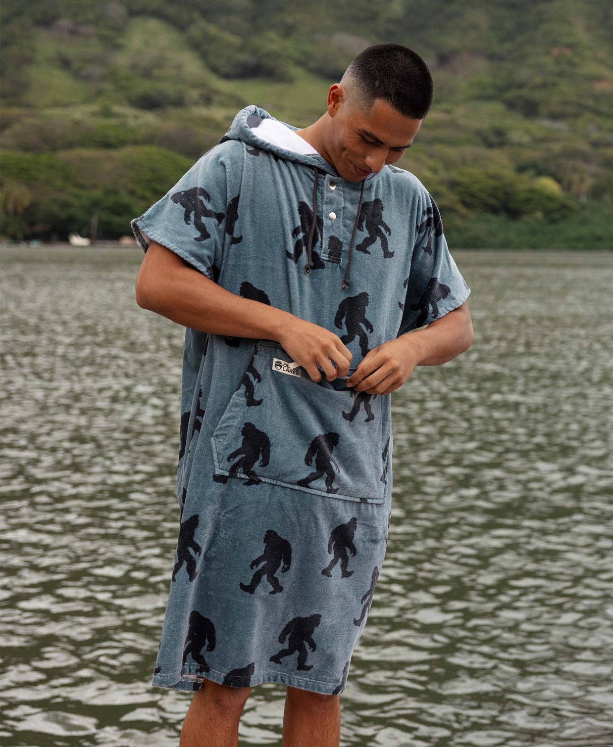 Person wearing a blue and grey bigfoot patterned towel poncho standing by a body of water.