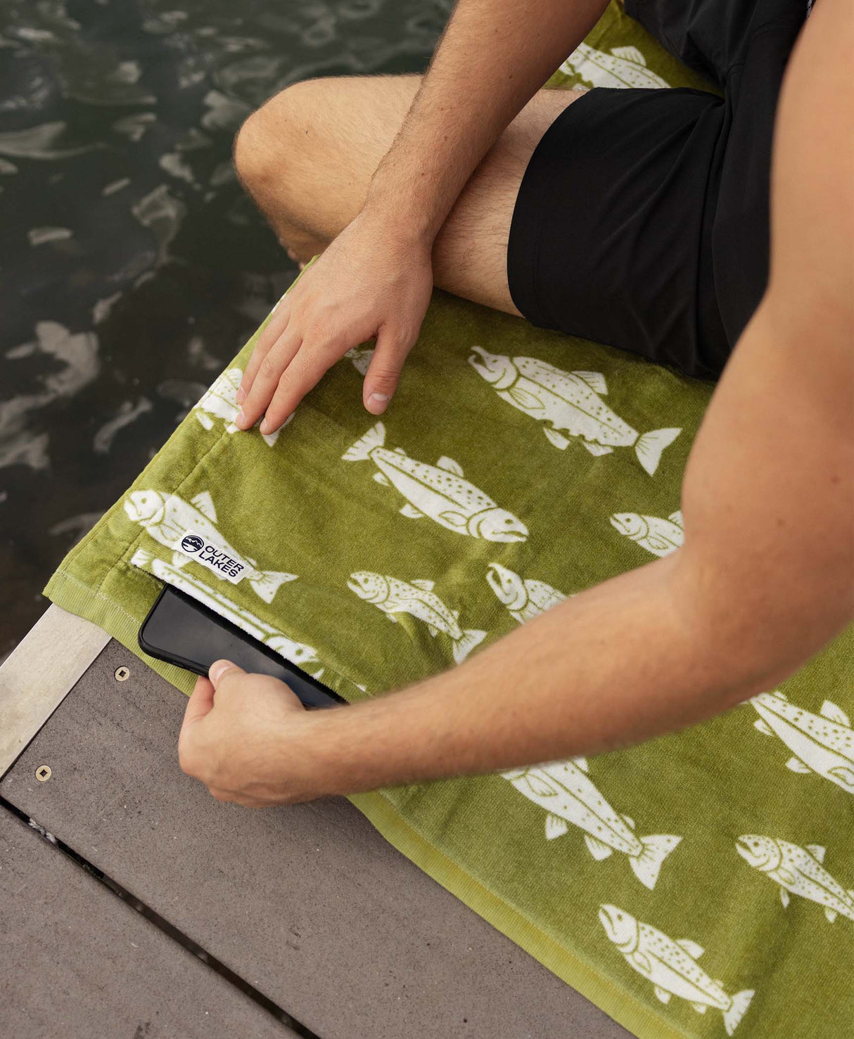 Person tucking phone into zipper pocket of a green fish-print towel by the water.