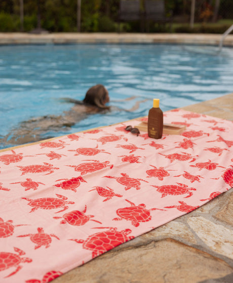Pink turtle towel with on a poolside with a person swimming in the background.