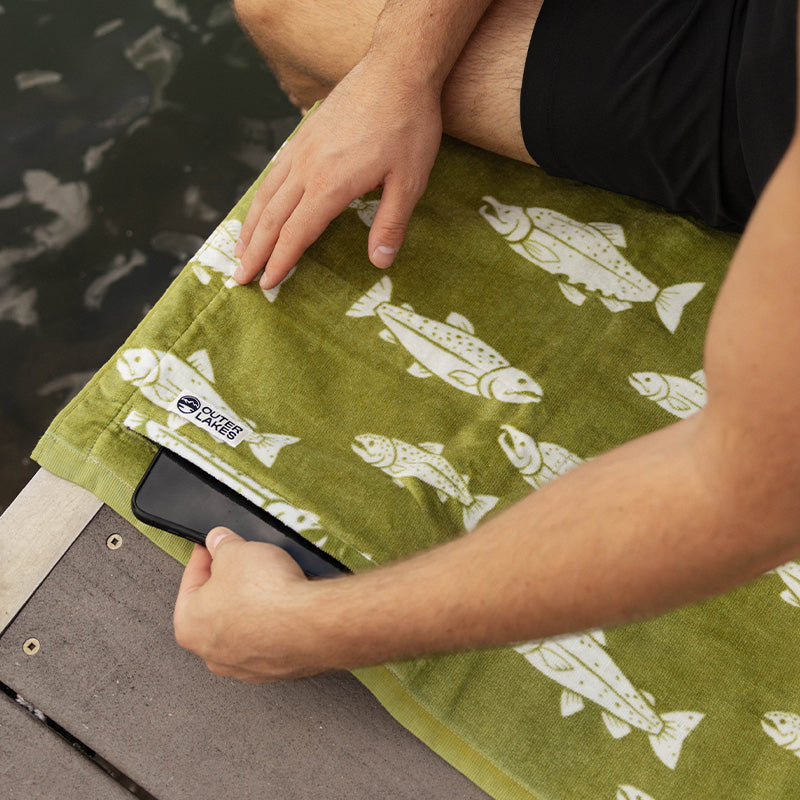 Person tucking phone into zipper pocket of a green fish-print towel by the water.