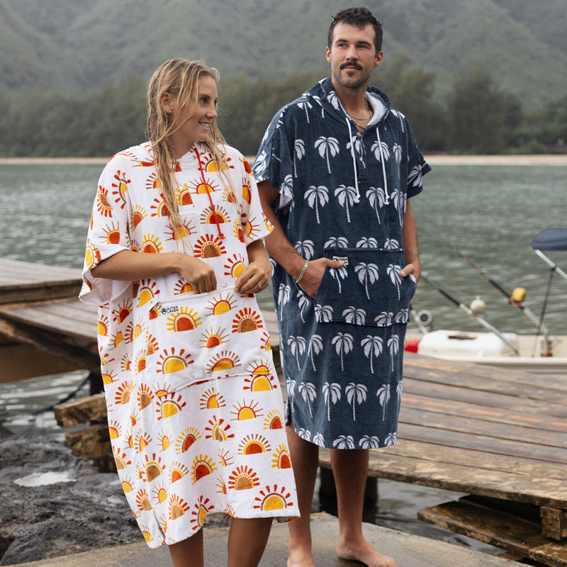 Two people in towel ponchos by a mountain lake — sun and palm tree patterns.