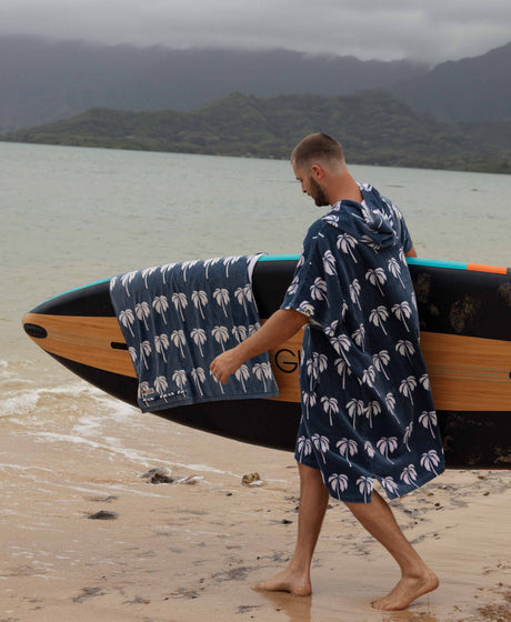 Man walking on a beach with a paddleboard covered by a towel with palm tree pattern