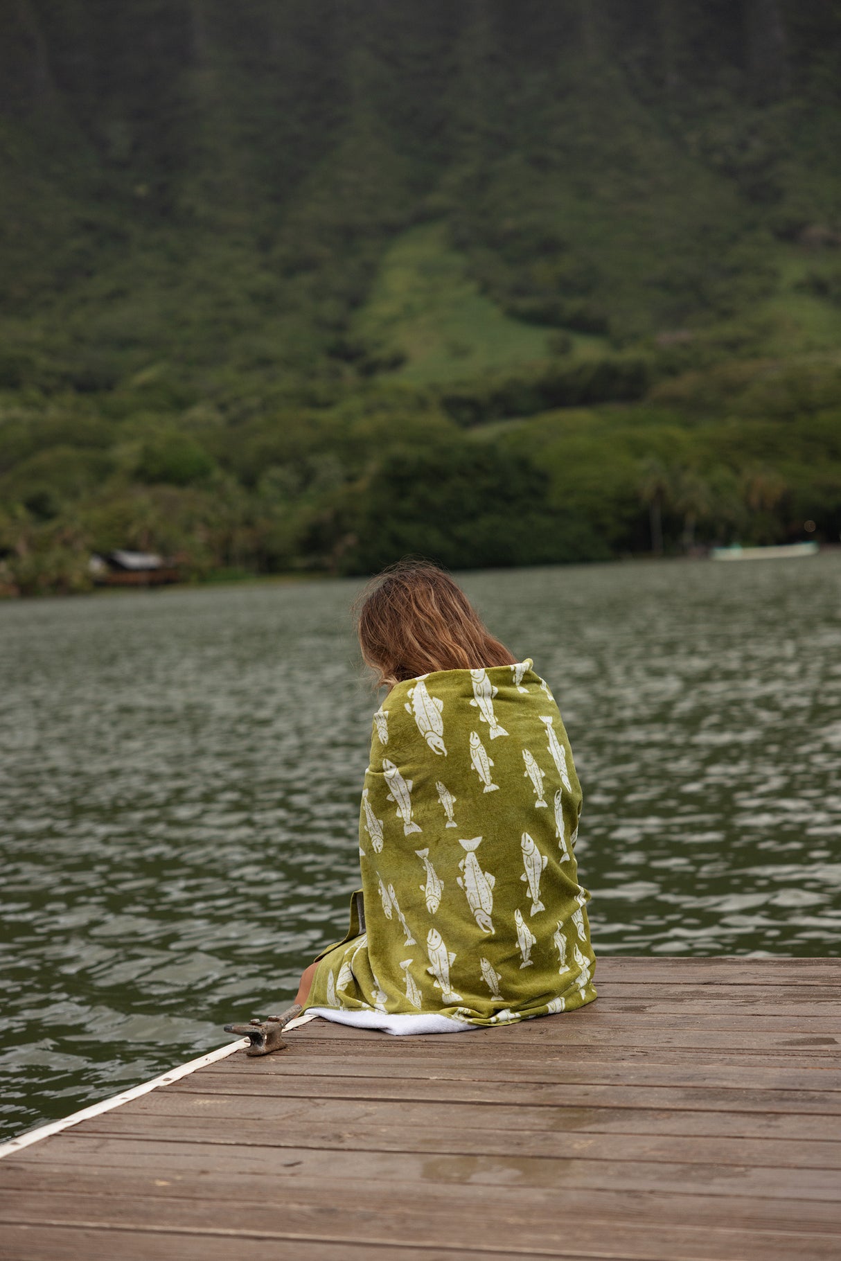 Person wrapped in a green and white patterned towel sitting on a wooden dock by a lake.