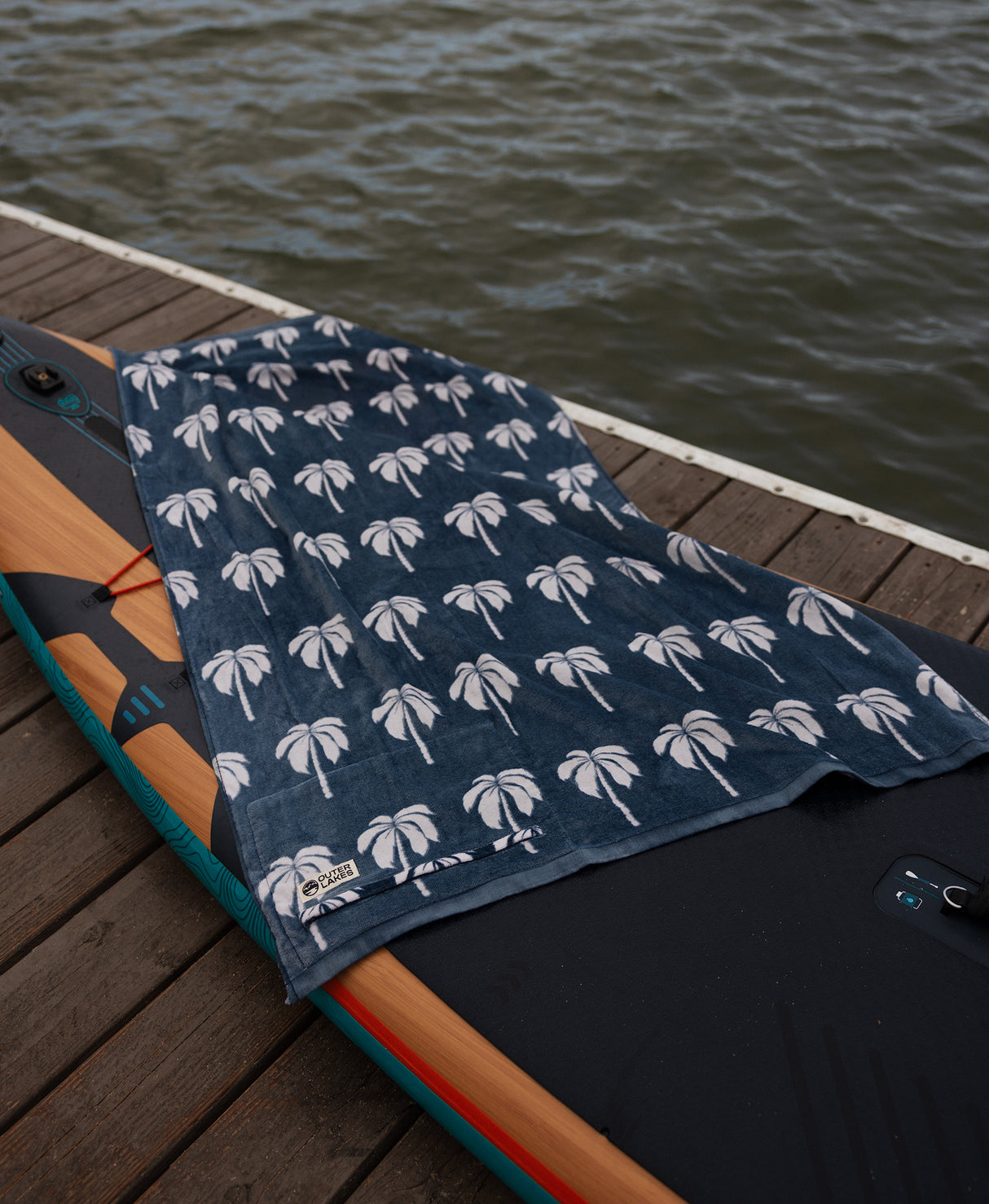 Navy towel with white palm tree pattern draped over a surfboard on a wooden dock by water.