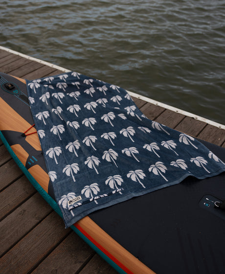 Navy towel with white palm tree pattern draped over a surfboard on a wooden dock by water.