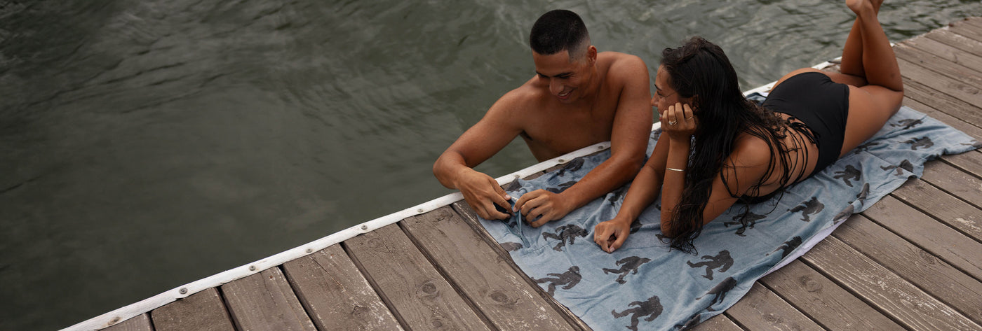 Two people at a dock, one in the water, one sitting on a Bigfoot-print towel.