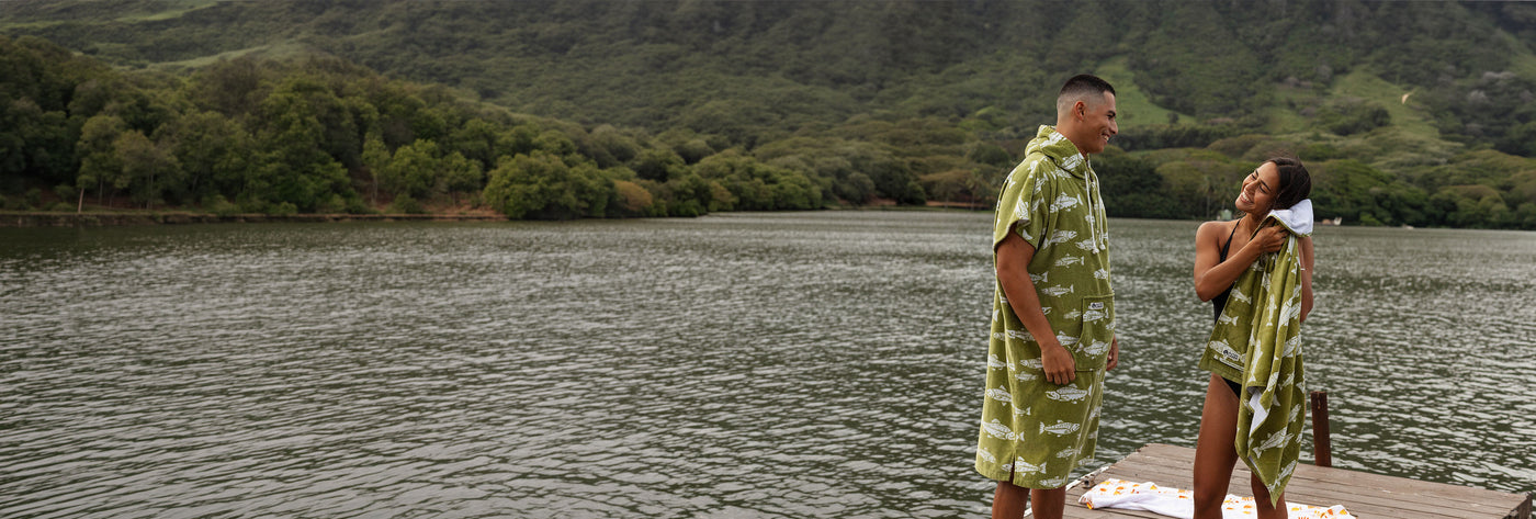 Two people in matching green fish-print towels on a dock with mountain backdrop.