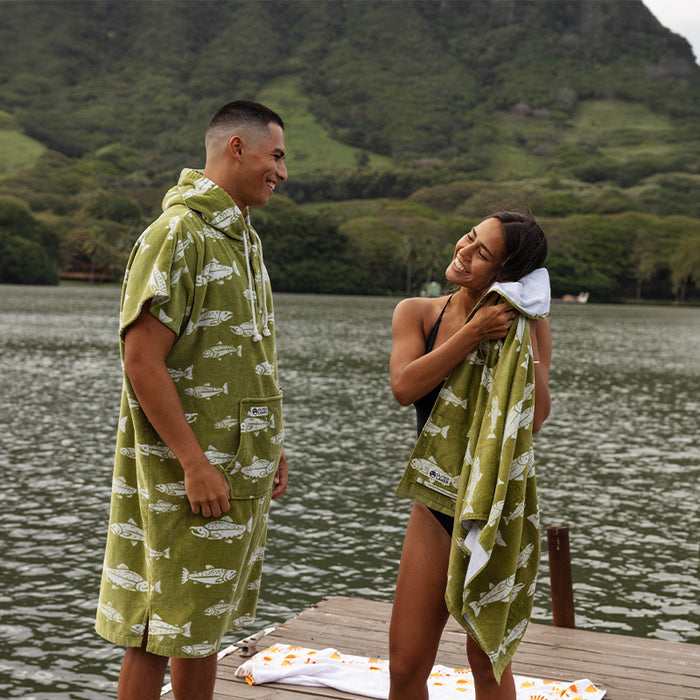 Two people in matching green fish-print towels on a dock with mountain backdrop.