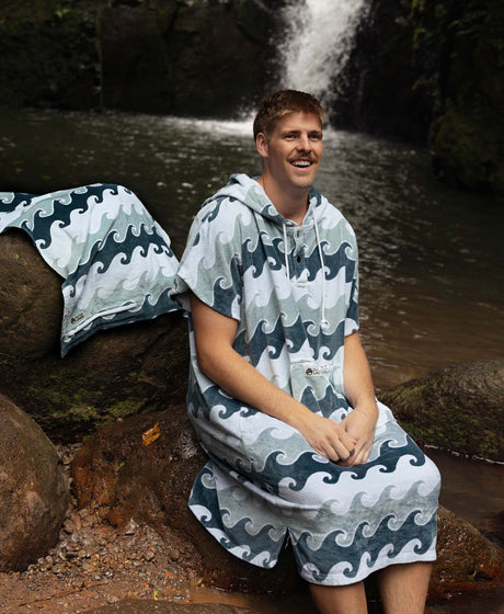 Man in wave-print towel poncho by a waterfall with matching towel beside him.