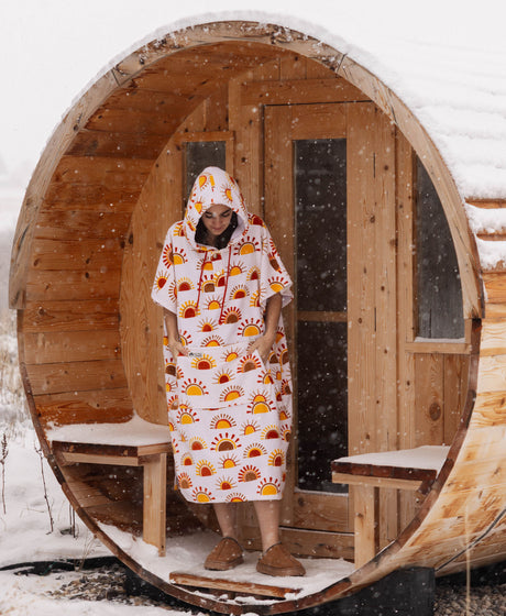 Person in sun-print towel poncho at the entrance of an outdoor sauna in the snow.