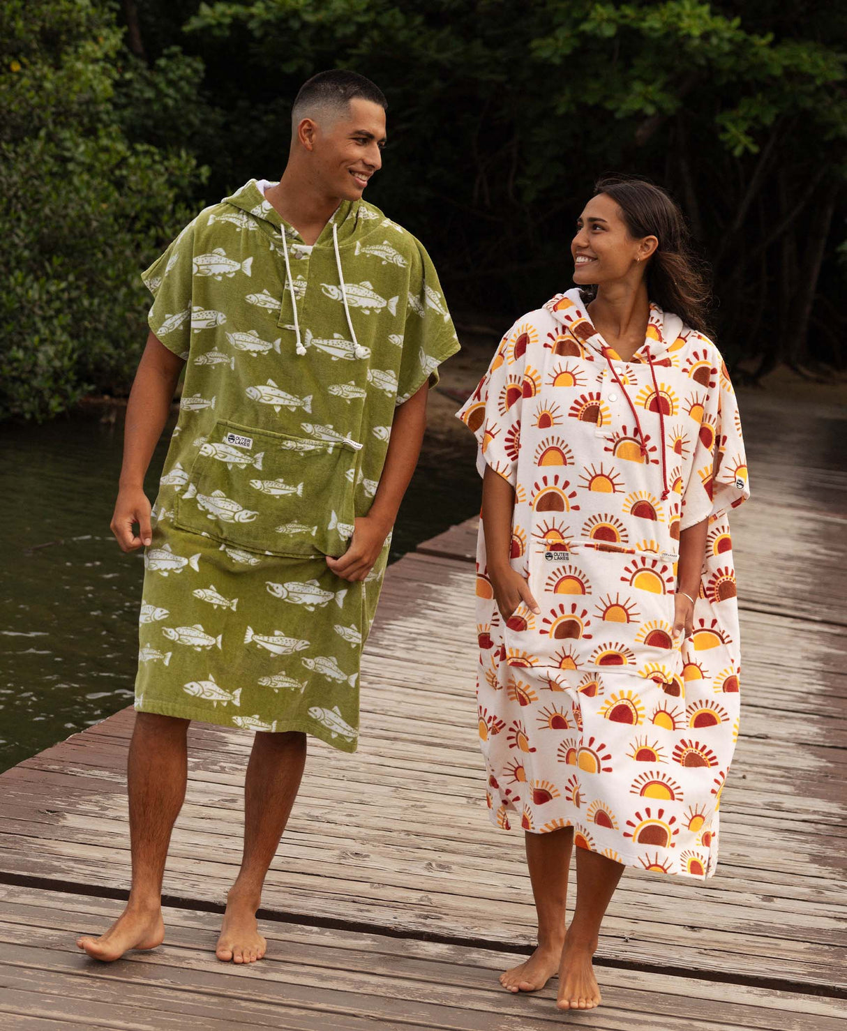 Two people wearing a fish patterned and sun patterned towel ponchos on a wooden dock.