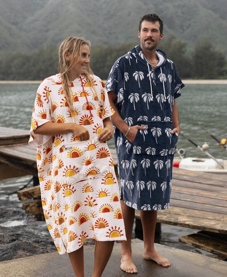 Two people in towel ponchos by a mountain lake — sun and palm tree patterns.