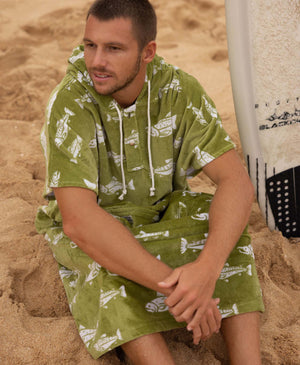 Man wearing a green fish patterned towel poncho sitting on sand with a surfboard behind him