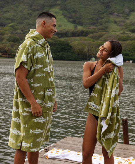 Two people in matching green fish-print towels on a dock with mountain backdrop.