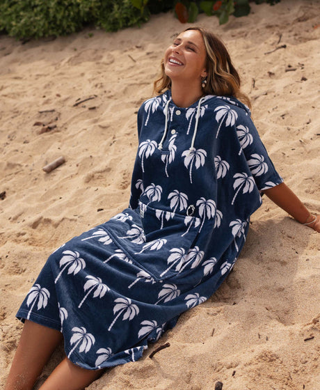 Woman sitting on sand wearing a navy blue towel poncho with white palm tree patterns