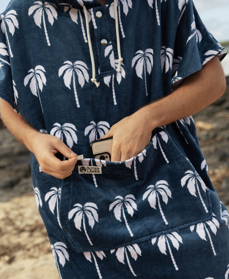 Person wearing a navy blue towel poncho with white palm tree pattern, putting a phone in zipper pocket