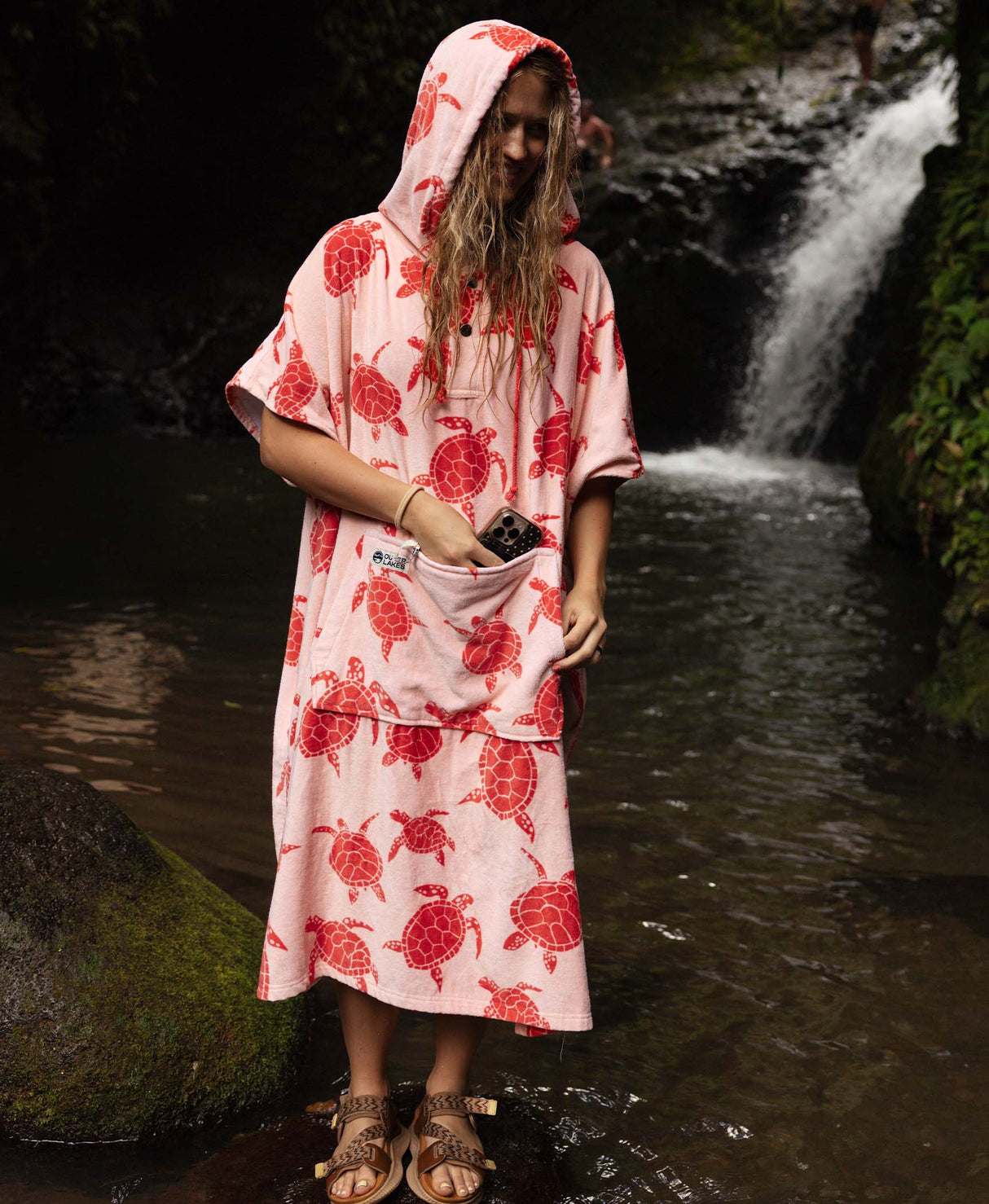 Woman in a pink turtle-print towel poncho standing in front of a waterfall.