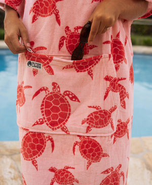 Person putting sunglasses in zipper pocket of pink turtle patterned towel poncho by a pool.