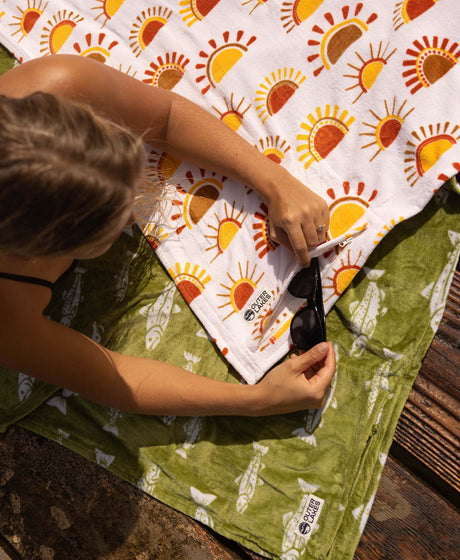 Person sitting on a green towel with a sun-patterned blanket, holding sunglasses.