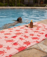 Pink turtle towel with on a poolside with a person swimming in the background.
