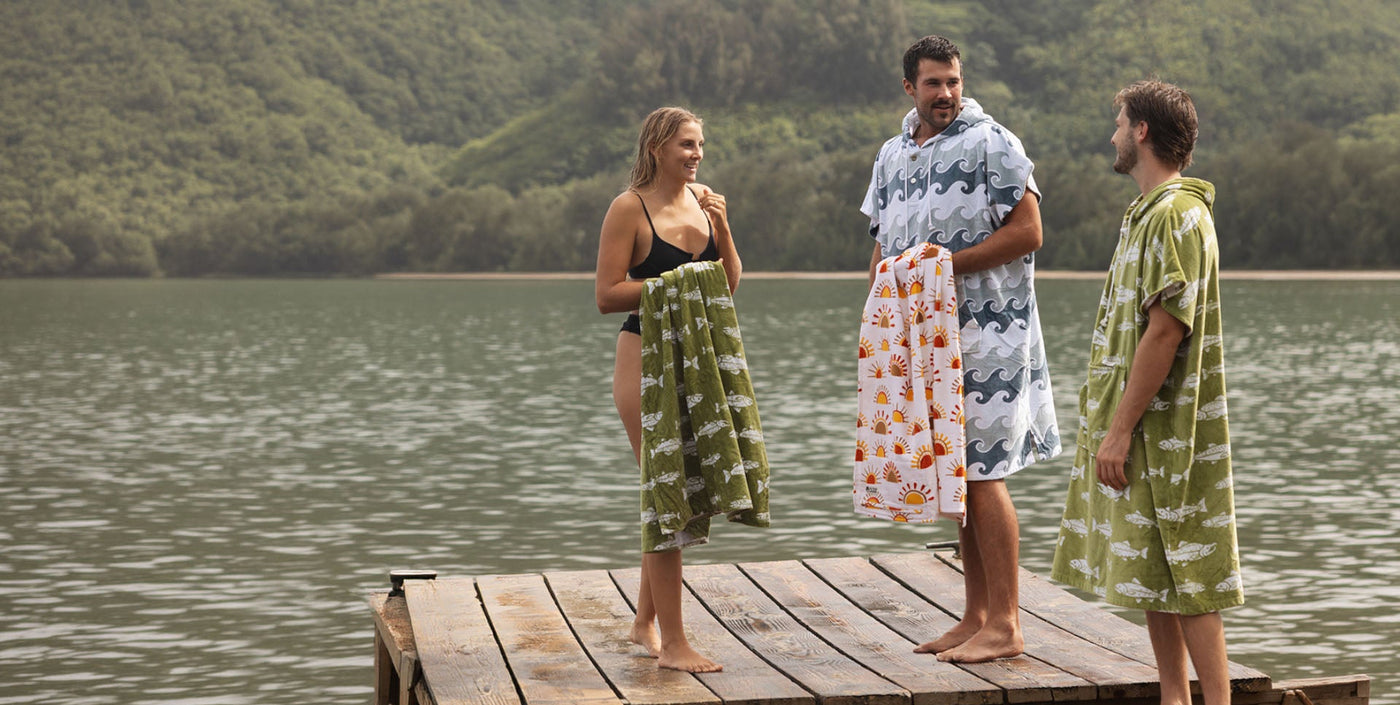 Three people on a dock, two in towel ponchos, one holding a towel.