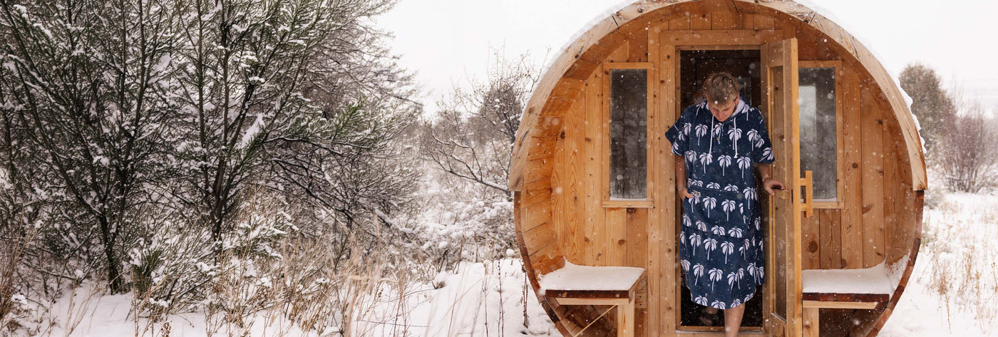 Person in a blue palm tree-print towel poncho inside a barrel sauna in the snow.