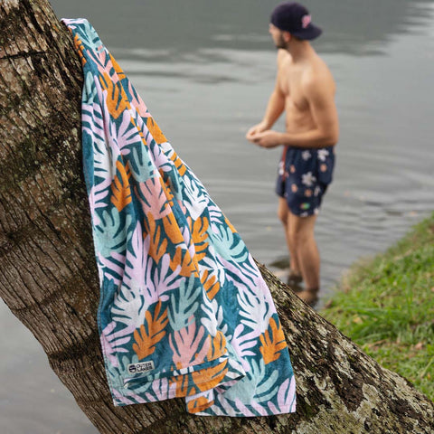 Coral patterned towel draped over a tree by a body of water with a person in swim shorts in the background.
