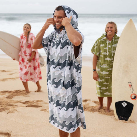 Person wearing a hooded towel poncho with wave pattern on a beach, with two other people in the background holding surfboards.