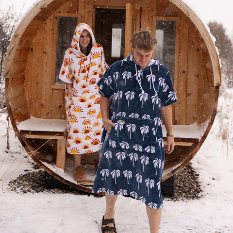 Two people wearing patterned hooded towel ponchos standing in front of a wooden sauna in the snow.