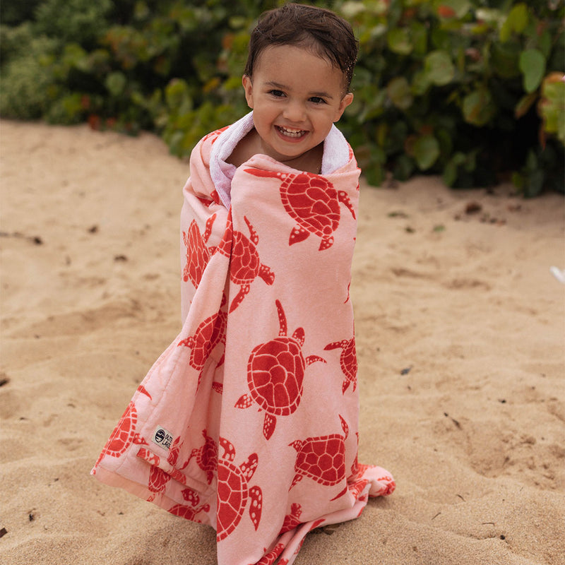 Child wrapped in a pink towel with dark pink turtle pattern on a sandy beach