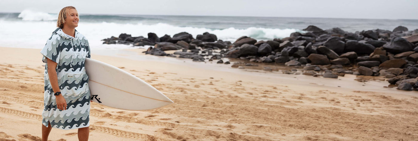 Person in a wave-print towel poncho holding a surfboard on a rocky beach.